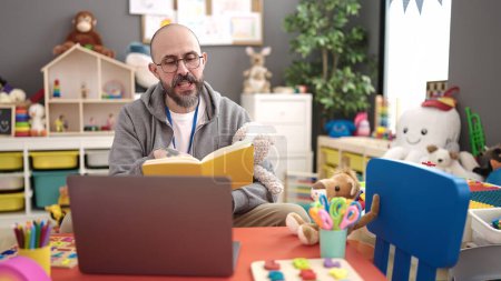 Photo for Young bald man preschool teacher reading story book on a video call at kindergarten - Royalty Free Image