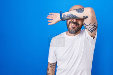 Téléchargez les photos : Homme hispanique avec des tatouages debout sur fond bleu couvrant les yeux avec le bras, l'air grave et triste. concept de non-visibilité, de dissimulation et de rejet - en image libre de droit