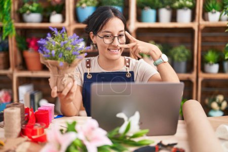 Photo for Young hispanic woman working at florist shop doing video call doing peace symbol with fingers over face, smiling cheerful showing victory - Royalty Free Image