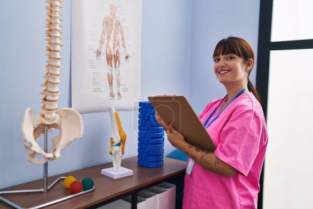 Photo for Young beautiful hispanic woman physiotherapist writing on document looking anatomical model at rehab clinic - Royalty Free Image