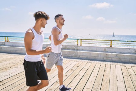 Téléchargez les photos : Deux hommes hispaniques couple sportif souriant confiant courir au bord de la mer - en image libre de droit