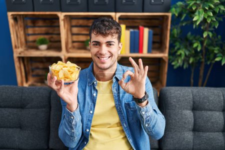 Téléchargez les photos : Jeune homme hispanique tenant des croustilles de pomme de terre faisant signe ok avec les doigts, sourire geste amical excellent symbole - en image libre de droit