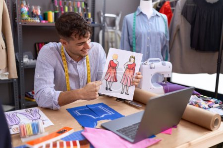 Téléchargez les photos : Jeune homme hispanique tailleur ayant appel vidéo montrant la conception de vêtements à l'atelier - en image libre de droit