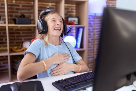 Téléchargez les photos : Jeune femme caucasienne jouant à des jeux vidéo portant des écouteurs souriant et riant fort à haute voix parce que drôle blague folle avec les mains sur le corps. - en image libre de droit