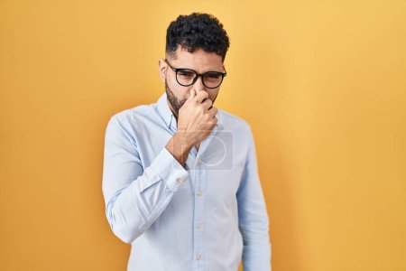 Téléchargez les photos : Homme hispanique avec la barbe debout sur fond jaune sentant quelque chose de puant et dégoûtant, odeur intolérable, retenant souffle avec les doigts sur le nez. mauvaise odeur - en image libre de droit
