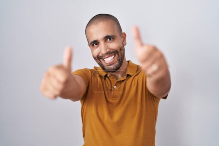 Foto de Hispanic man with beard standing over white background approving doing positive gesture with hand, thumbs up smiling and happy for success. winner gesture. - Imagen libre de derechos