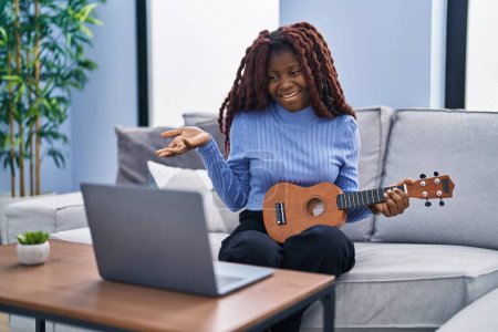 Photo for African woman playing ukulele at home doing video call celebrating achievement with happy smile and winner expression with raised hand - Royalty Free Image