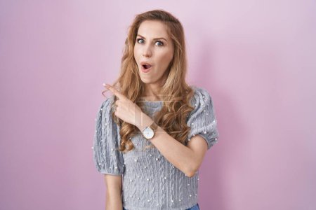 Photo for Beautiful blonde woman standing over pink background surprised pointing with finger to the side, open mouth amazed expression. - Royalty Free Image