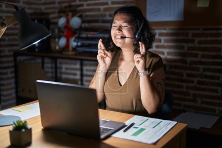 Téléchargez les photos : Young hispanic woman working at the office at night gesturing finger crossed smiling with hope and eyes closed. luck and superstitious concept. - en image libre de droit