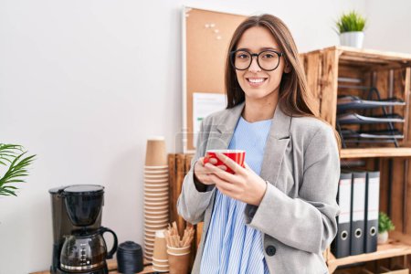 Foto de Joven hermosa mujer hispana trabajadora de negocios sonriendo confiada bebiendo café en la oficina - Imagen libre de derechos