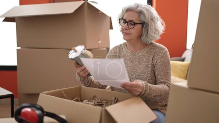 Téléchargez les photos : Middle age woman with grey hair unpacking cardboard box reading document at new home - en image libre de droit
