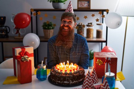 Foto de Hombre caucásico con barba larga celebrando cumpleaños sosteniendo gran pastel de chocolate mirando positiva y feliz de pie y sonriendo con una sonrisa confiada mostrando los dientes - Imagen libre de derechos