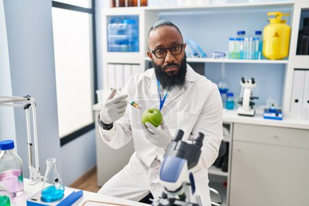 Photo for African american man working at scientist laboratory with apple relaxed with serious expression on face. simple and natural looking at the camera. - Royalty Free Image