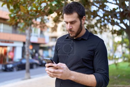 Téléchargez les photos : Jeune homme hispanique utilisant un smartphone avec une expression détendue au parc - en image libre de droit