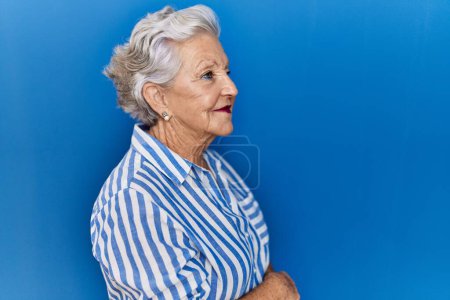 Téléchargez les photos : Femme âgée avec les cheveux gris debout sur fond bleu regardant vers le côté, pose de profil relax avec le visage naturel avec sourire confiant. - en image libre de droit