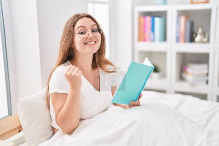 Photo for Young caucasian woman sitting on the bed at home reading a book screaming proud, celebrating victory and success very excited with raised arms - Royalty Free Image