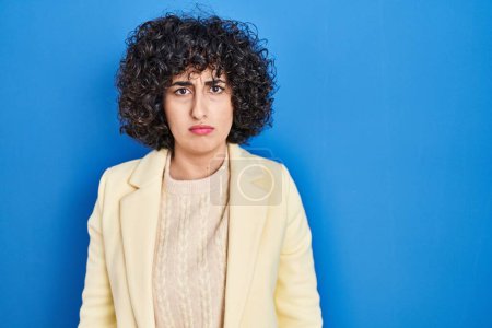 Photo for Young brunette woman with curly hair standing over blue background skeptic and nervous, frowning upset because of problem. negative person. - Royalty Free Image