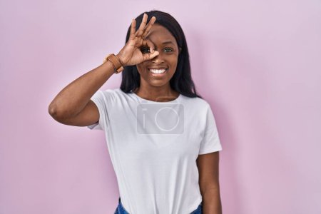 Foto de Mujer joven africana con camiseta blanca casual haciendo buen gesto con la mano sonriente, ojos mirando a través de los dedos con cara feliz. - Imagen libre de derechos