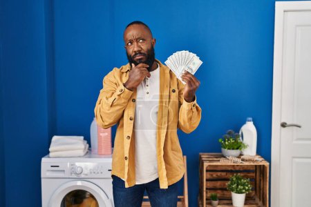 Téléchargez les photos : African american man holding money at laundry room serious face thinking about question with hand on chin, thoughtful about confusing idea - en image libre de droit
