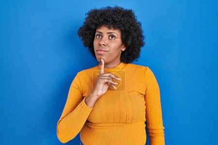Foto de Mujer negra con el pelo rizado de pie sobre fondo azul pensando concentrado en la duda con el dedo en la barbilla y mirando hacia arriba preguntándose - Imagen libre de derechos