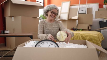 Photo for Middle age woman with grey hair smiling confident unpacking cardboard box at new home - Royalty Free Image