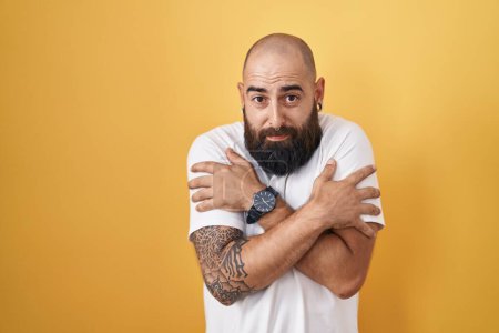 Téléchargez les photos : Young hispanic man with beard and tattoos standing over yellow background shaking and freezing for winter cold with sad and shock expression on face - en image libre de droit