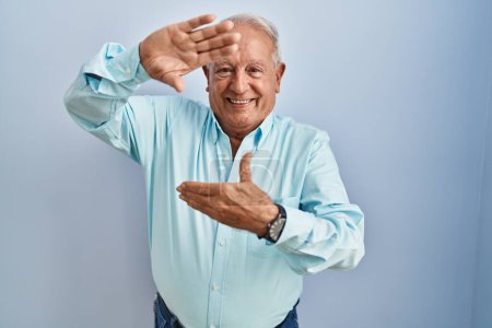 Foto de Hombre mayor con el pelo gris de pie sobre fondo azul sonriendo haciendo marco con las manos y los dedos con la cara feliz. concepto de creatividad y fotografía. - Imagen libre de derechos