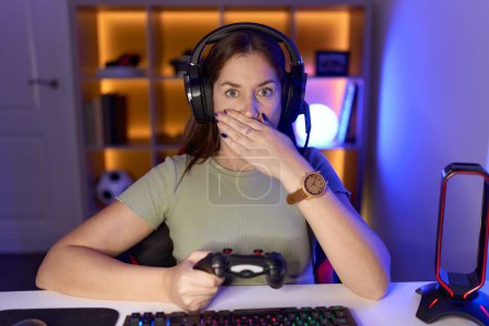 Photo for Beautiful brunette woman playing video games wearing headphones shocked covering mouth with hands for mistake. secret concept. - Royalty Free Image