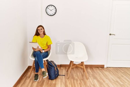 Téléchargez les photos : Jeune étudiante tenant un cahier assis sur une chaise dans la salle d'attente - en image libre de droit
