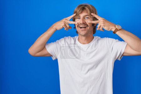 Foto de Middle age man standing over blue background doing peace symbol with fingers over face, smiling cheerful showing victory - Imagen libre de derechos