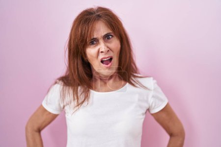 Foto de Mujer de mediana edad de pie sobre fondo rosa en la cara de choque, mirando escéptico y sarcástico, sorprendido con la boca abierta - Imagen libre de derechos