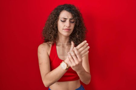 Foto de Mujer hispana con el pelo rizado de pie sobre fondo rojo sufriendo dolor en las manos y los dedos, inflamación de la artritis - Imagen libre de derechos