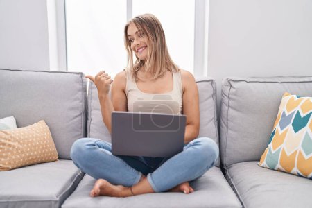 Téléchargez les photos : Blonde caucasian woman using laptop at home sitting on the sofa pointing thumb up to the side smiling happy with open mouth - en image libre de droit