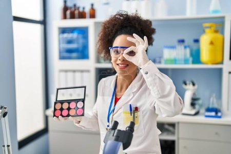 Foto de Mujer afroamericana joven que trabaja en el laboratorio científico haciendo maquillaje sonriendo feliz haciendo signo de ok con la mano en el ojo mirando a través de los dedos - Imagen libre de derechos
