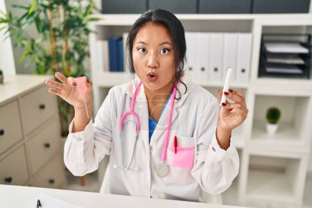 Téléchargez les photos : Young asian doctor woman holding menstrual cup and tampon afraid and shocked with surprise and amazed expression, fear and excited face. - en image libre de droit