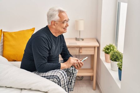 Téléchargez les photos : Homme âgé utilisant smartphone assis sur le lit dans la chambre - en image libre de droit