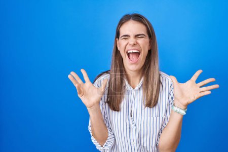 Photo for Hispanic young woman standing over blue background celebrating mad and crazy for success with arms raised and closed eyes screaming excited. winner concept - Royalty Free Image