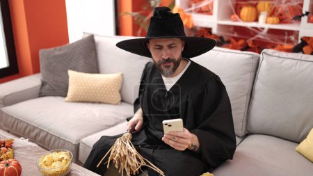 Téléchargez les photos : Jeune homme chauve portant un costume de sorcier à l'aide d'un smartphone tenant un balai à la maison - en image libre de droit