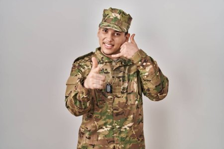 Téléchargez les photos : Jeune homme arabe portant un uniforme de camouflage de l'armée souriant faisant parler au téléphone geste et vous pointant du doigt. Appelle-moi. - en image libre de droit