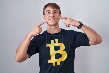 Téléchargez les photos : Homme blond caucasien portant Bitcoin t-shirt souriant gai montrant et pointant avec les doigts dents et la bouche. concept de santé dentaire. - en image libre de droit