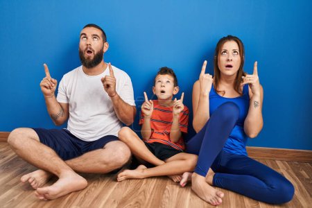 Foto de Familia de tres sentados en el suelo en casa asombrados y sorprendidos mirando hacia arriba y señalando con los dedos y los brazos levantados. - Imagen libre de derechos