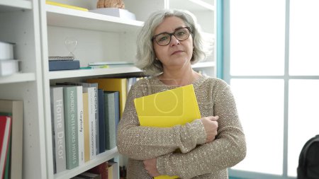 Foto de Middle age woman with grey hair teacher teaching maths lesson holding book at university classroom - Imagen libre de derechos