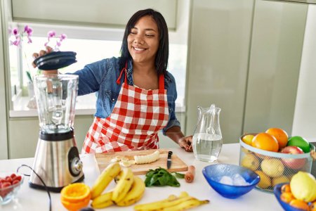 Téléchargez les photos : Femme brune hispanique préparant un smoothie aux fruits à la cuisine - en image libre de droit