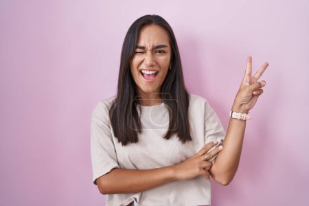 Photo for Young hispanic woman standing over pink background smiling with happy face winking at the camera doing victory sign with fingers. number two. - Royalty Free Image