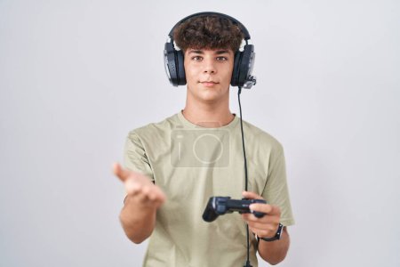 Foto de Adolescente hispana jugando videojuego sosteniendo controlador sonriente alegre ofreciendo mano de palma dando asistencia y aceptación. - Imagen libre de derechos