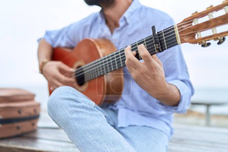 Téléchargez les photos : Young hispanic man musician playing classical guitar sitting on bench at seaside - en image libre de droit