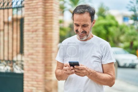 Téléchargez les photos : Moyen Age homme souriant confiant en utilisant smartphone à la rue - en image libre de droit