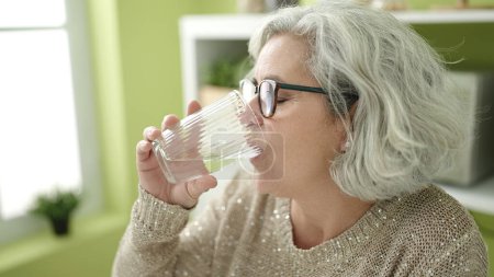 Foto de Mujer de mediana edad con pelo gris bebiendo vaso de agua sentado en la mesa en casa - Imagen libre de derechos