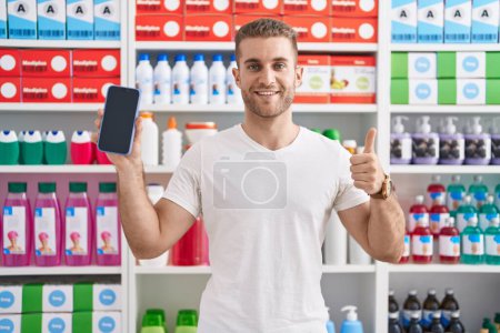 Téléchargez les photos : Jeune homme caucasien travaillant à la pharmacie pharmacie montrant écran smartphone souriant heureux et positif, pouce levé faire excellent et signe d'approbation - en image libre de droit