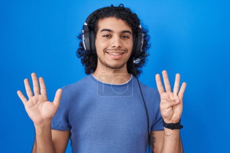 Téléchargez les photos : Homme hispanique aux cheveux bouclés écoutant de la musique à l'aide d'écouteurs montrant et pointant vers le haut avec les doigts numéro neuf tout en souriant confiant et heureux. - en image libre de droit